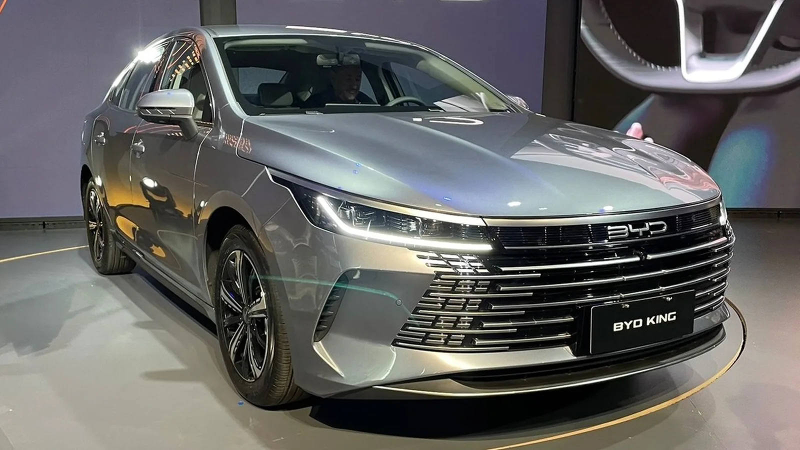 BYD King azul metálico em perfil lateral, carregando em estação moderna. Autonomia de 600 km em destaque.