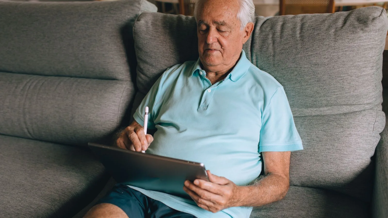Elderly man in blue polo shirt using a tablet while lounging on a comfortable sofa.