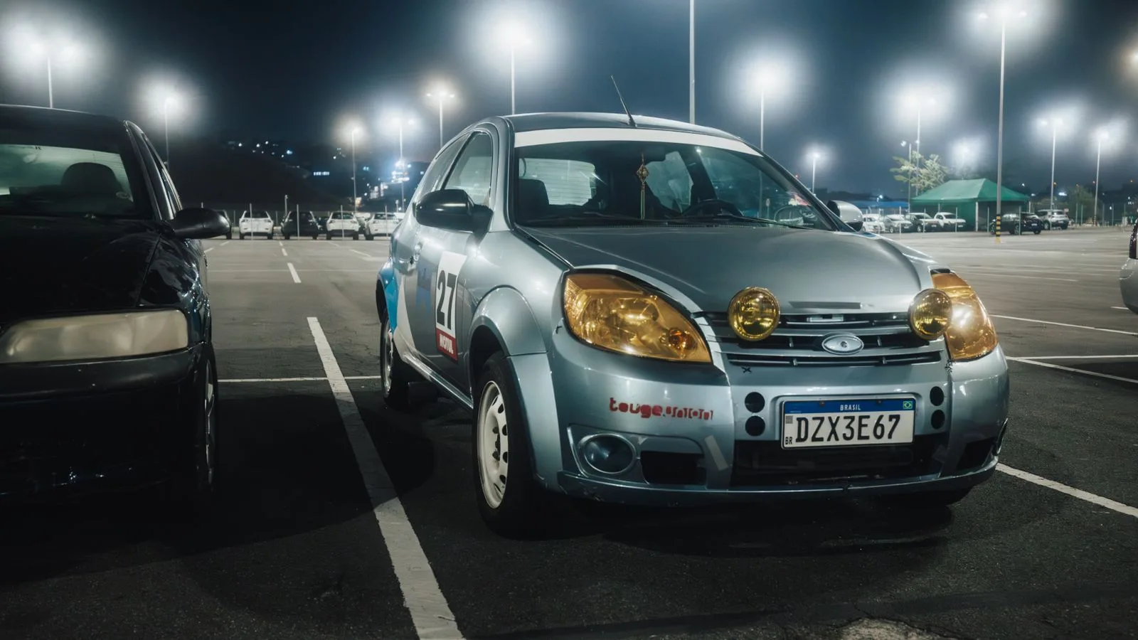 Silver car with yellow headlights in a lit parking lot at night.