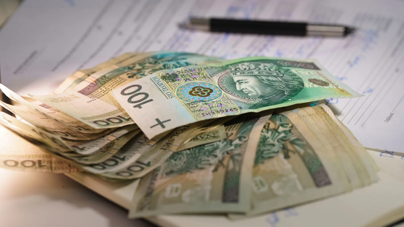 Stack of Polish zloty banknotes on financial documents with a pen, indicating monetary transactions in an office setting.