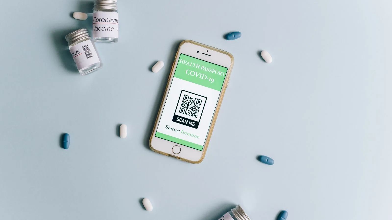 Image of a smartphone displaying a health passport alongside COVID-19 vaccine vials and pills on a light blue background.
