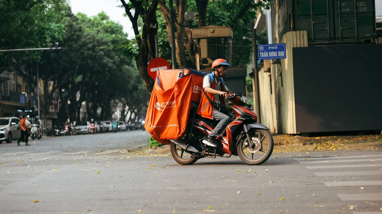 A delivery driver transports goods on a motorcycle in a bustling city street scene.