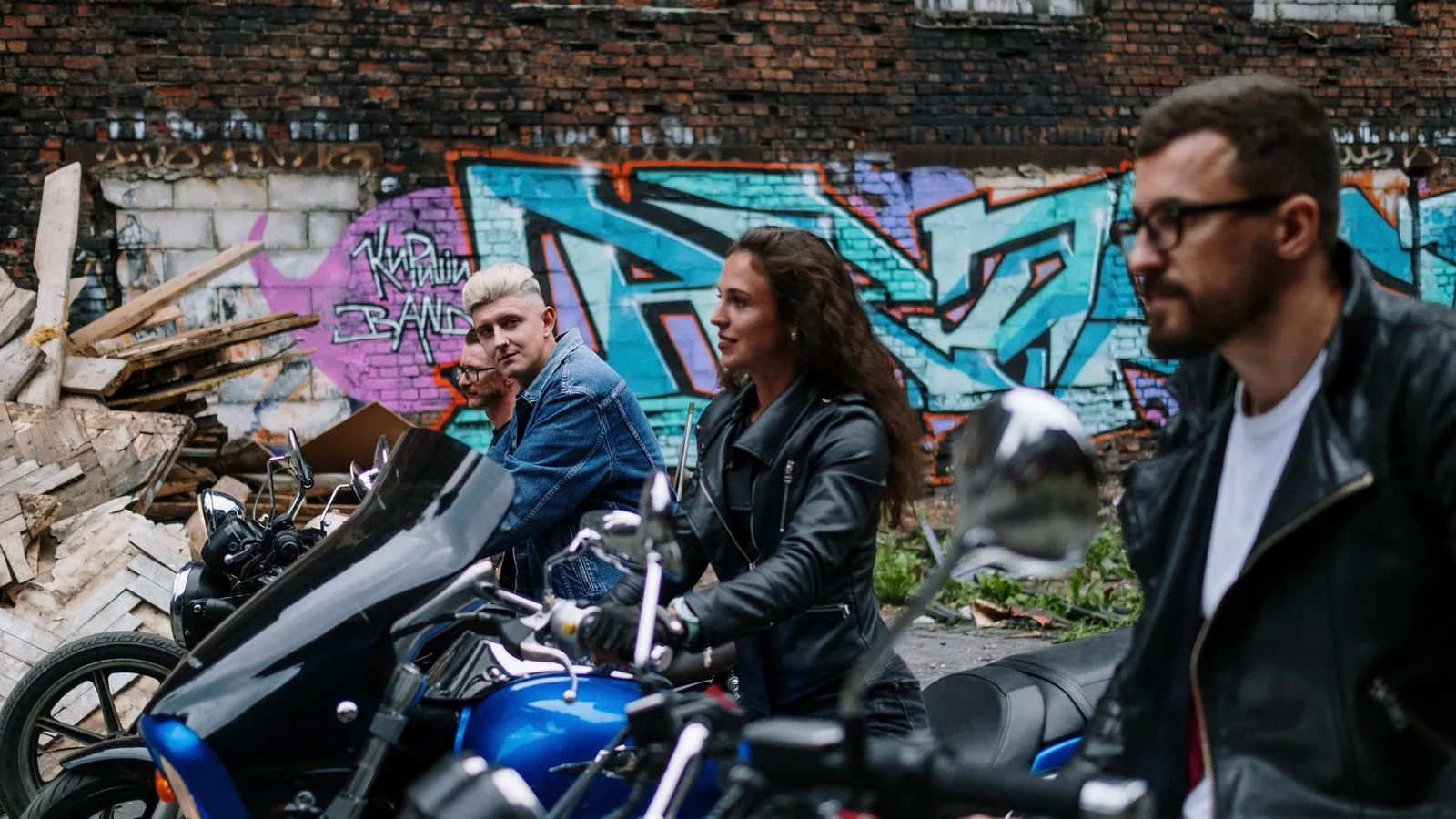 Three bikers with motorcycles against a backdrop of urban graffiti and a gritty brick wall.