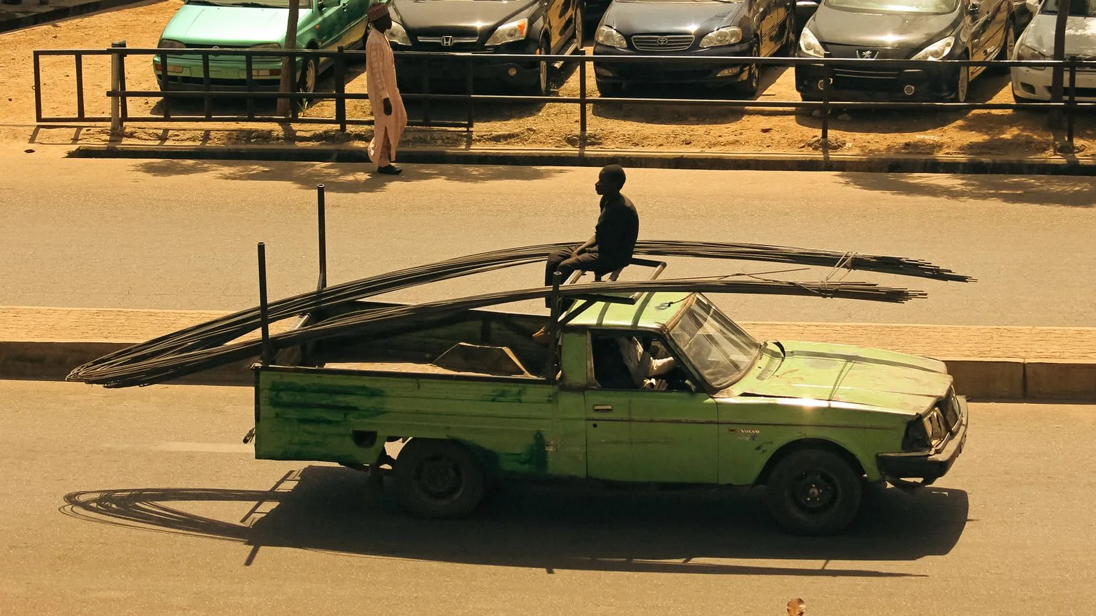 A vintage green pickup truck carrying metal rods with a man sitting on them, captured from above on a sunny day.