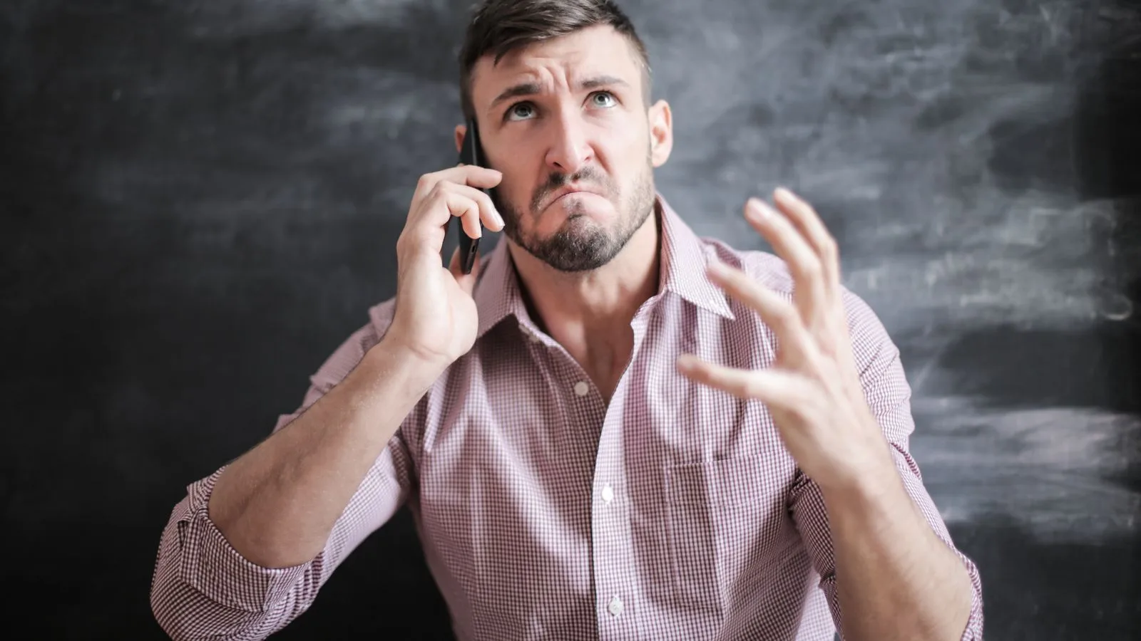 Adult man in pink dress shirt looking stressed while talking on a smartphone indoors.