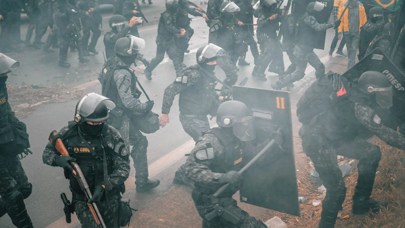 Riot police with shields confront protesters in a smoky urban setting.