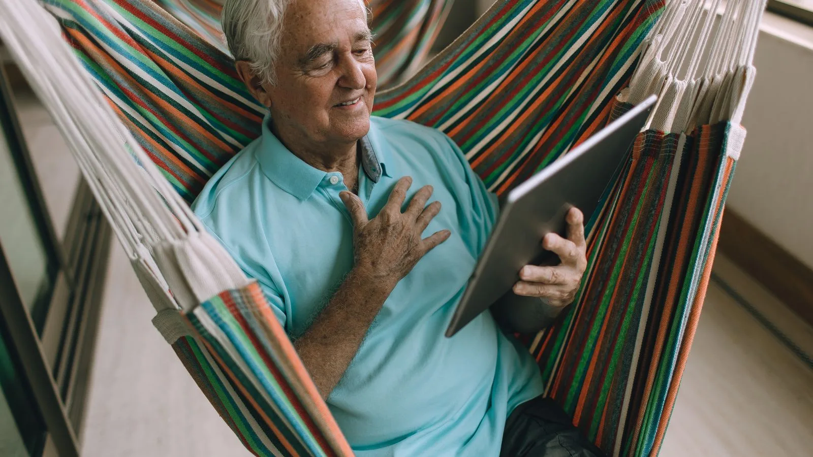 Elderly man enjoys a tablet while sitting comfortably in a colorful hammock indoors.