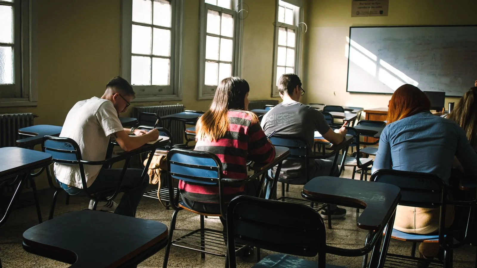 A group of students focused on writing in a sunlit classroom with desks and windows.