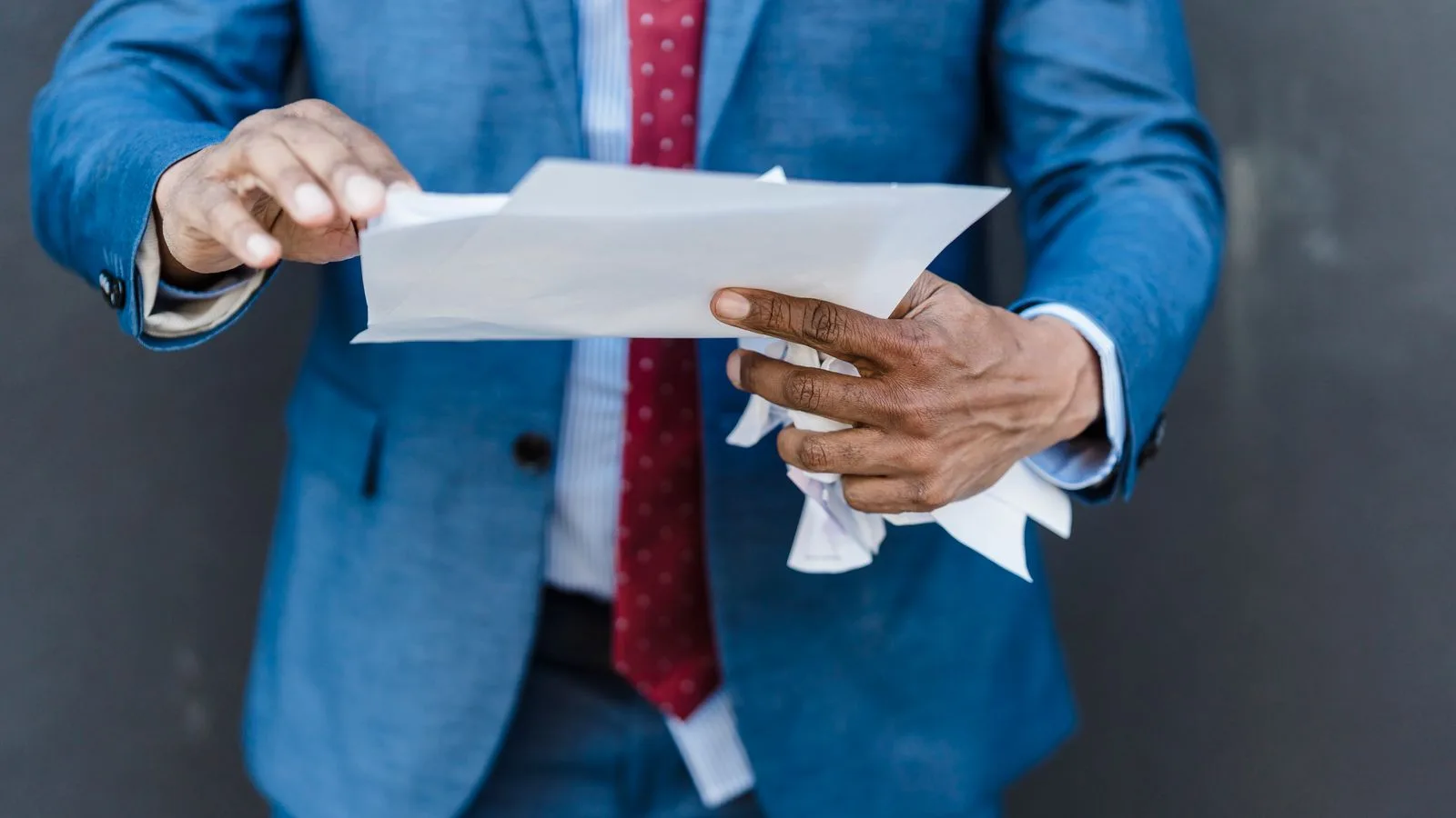 Crop anonymous businessman in classy suit with documents and crumpled paper in hands standing on gray background in city after hard workday