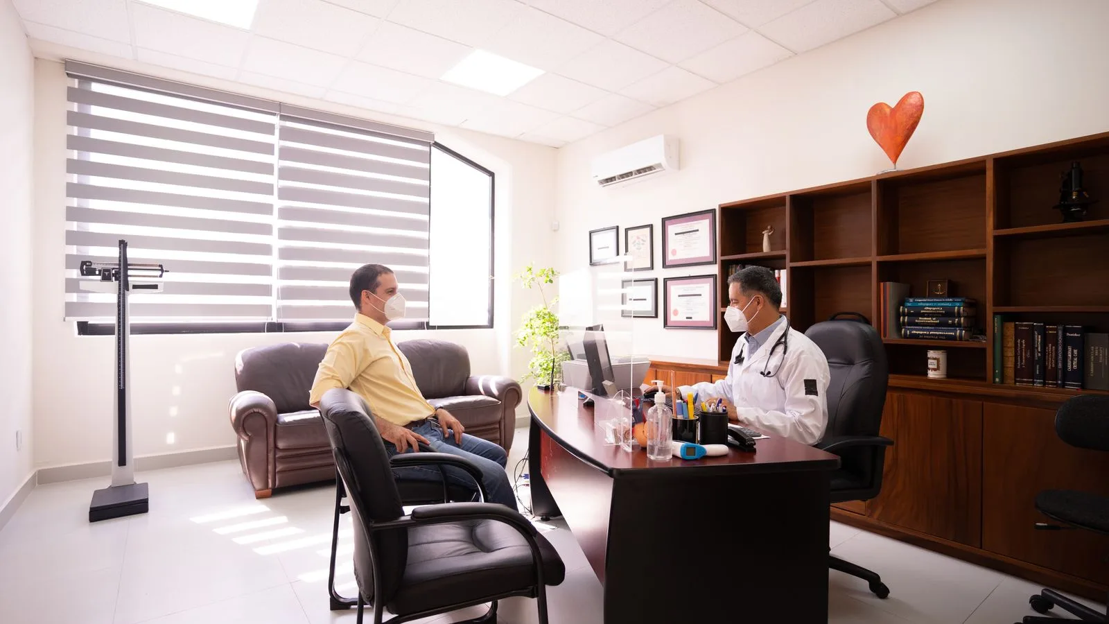 A patient consults with a masked doctor in a well-lit, modern office. Safety measures in place.