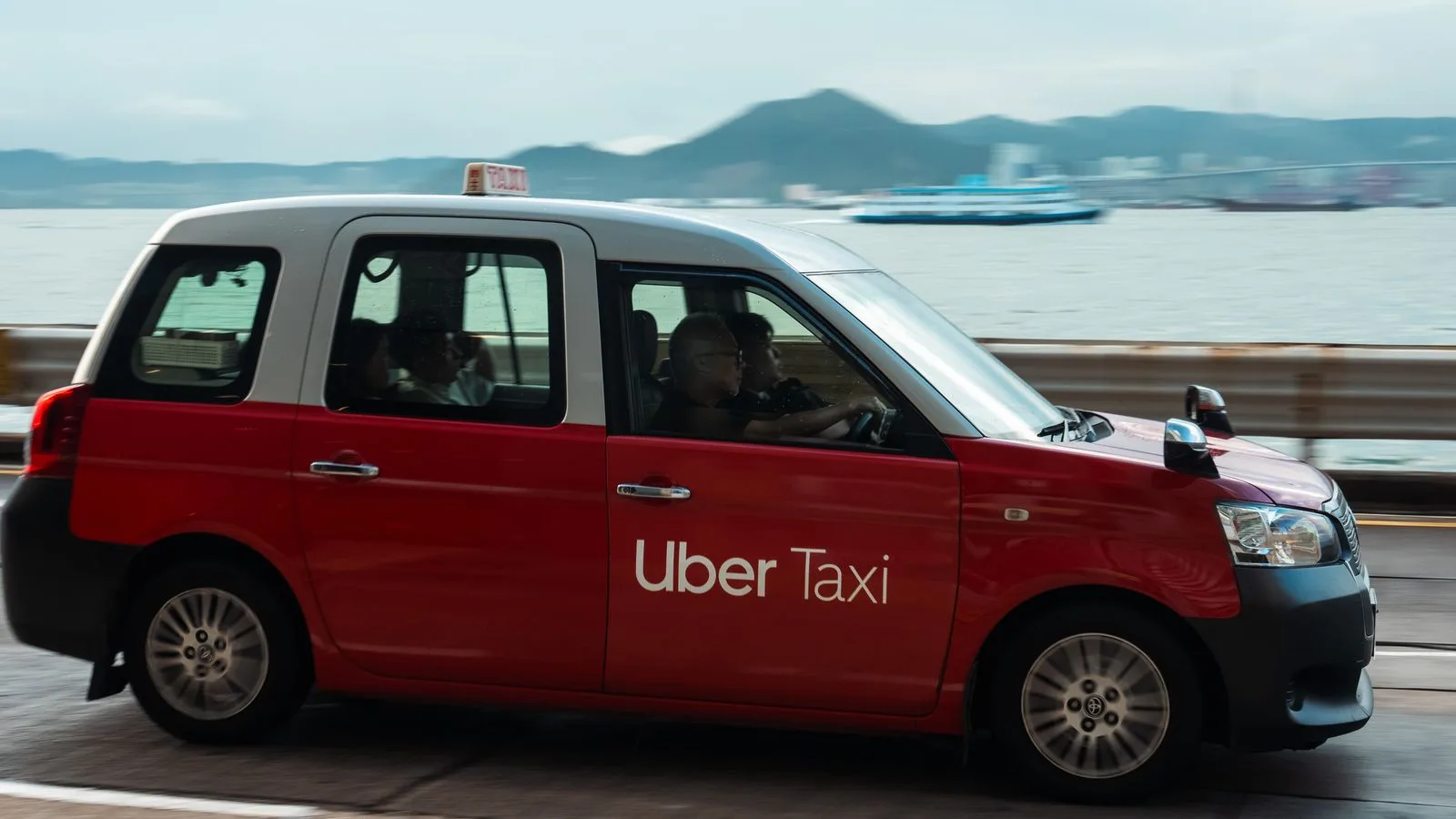 Red Uber taxi driving along a waterfront with a serene view of the sea and distant mountains.