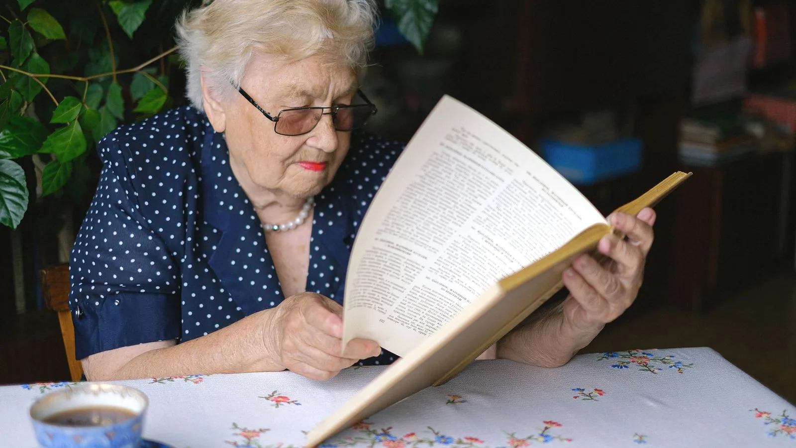Serious female pensioner in eyeglasses reading book while sitting at table with cup of hot drink in room on blurred background