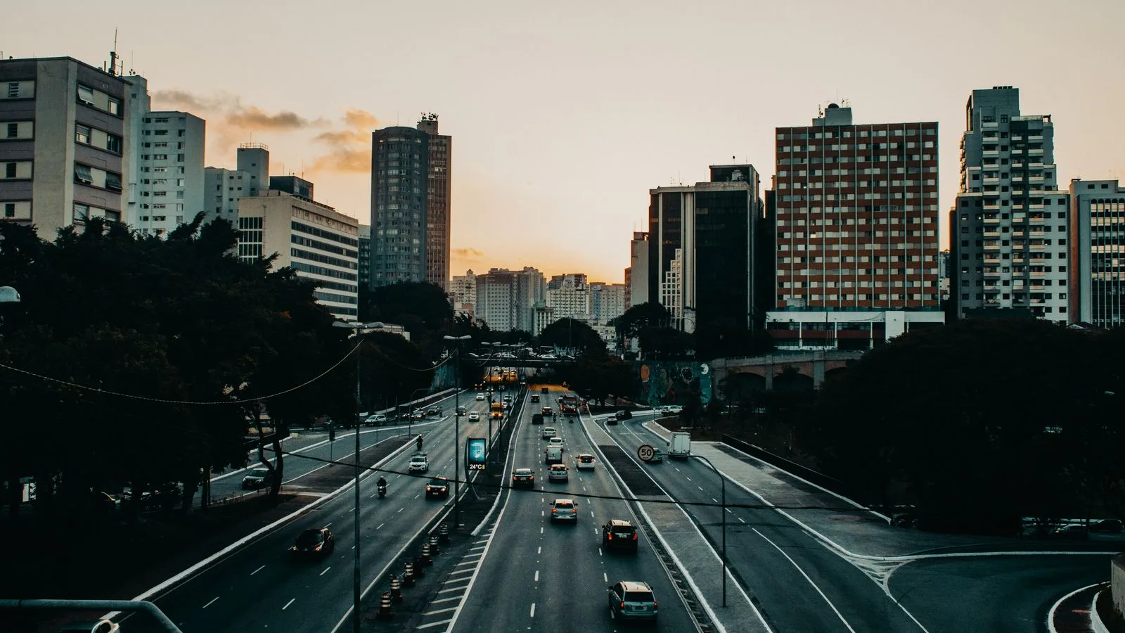 Various vehicles riding along asphalt road near modern buildings in city district against sunset sky