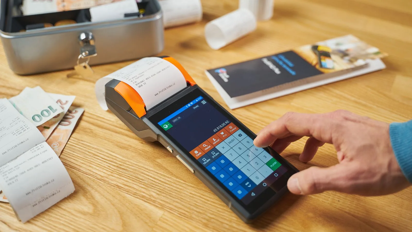 Person using a digital payment terminal on a wooden desk with cash and receipts visible.