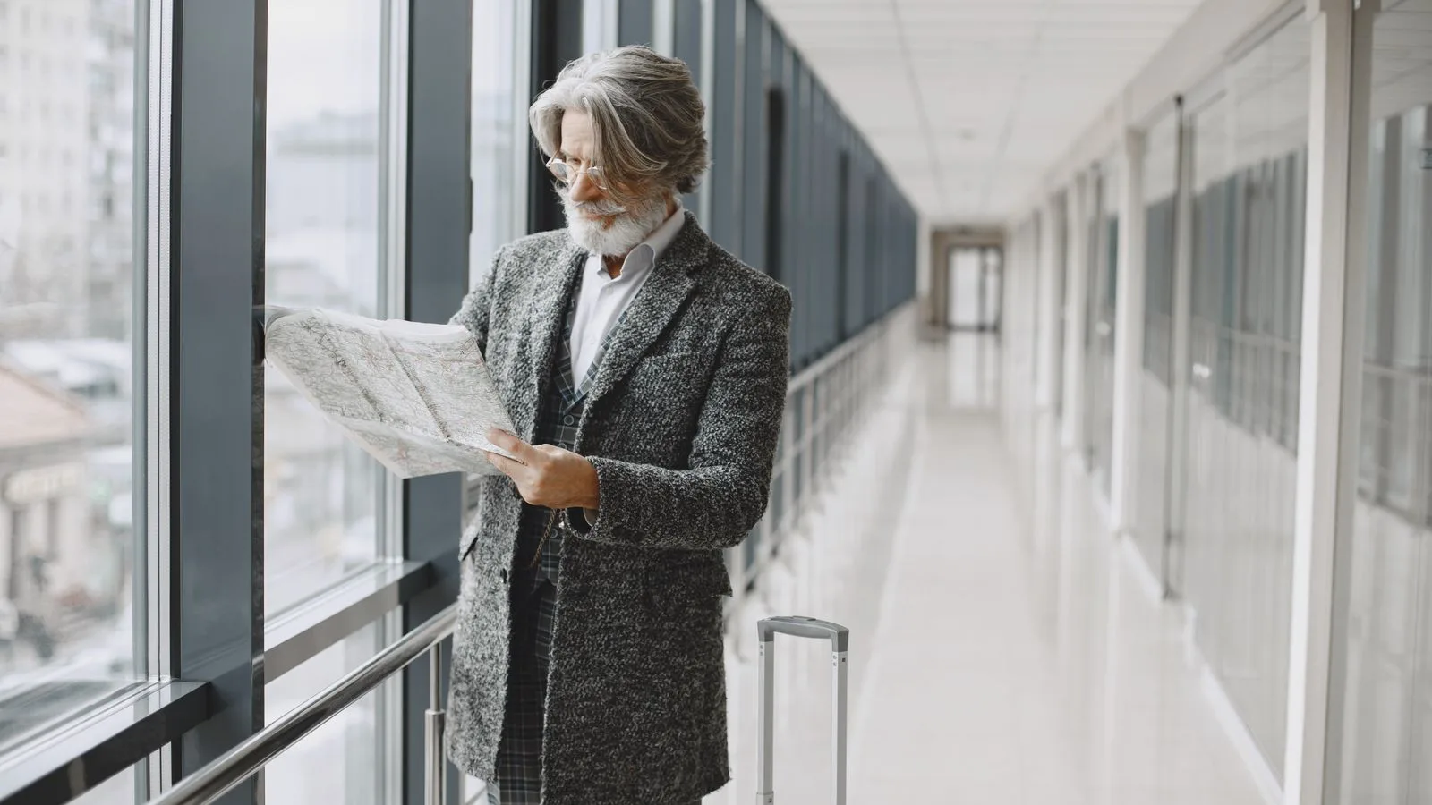 Senior man with beard reads map by glass windows in modern hallway with luggage.