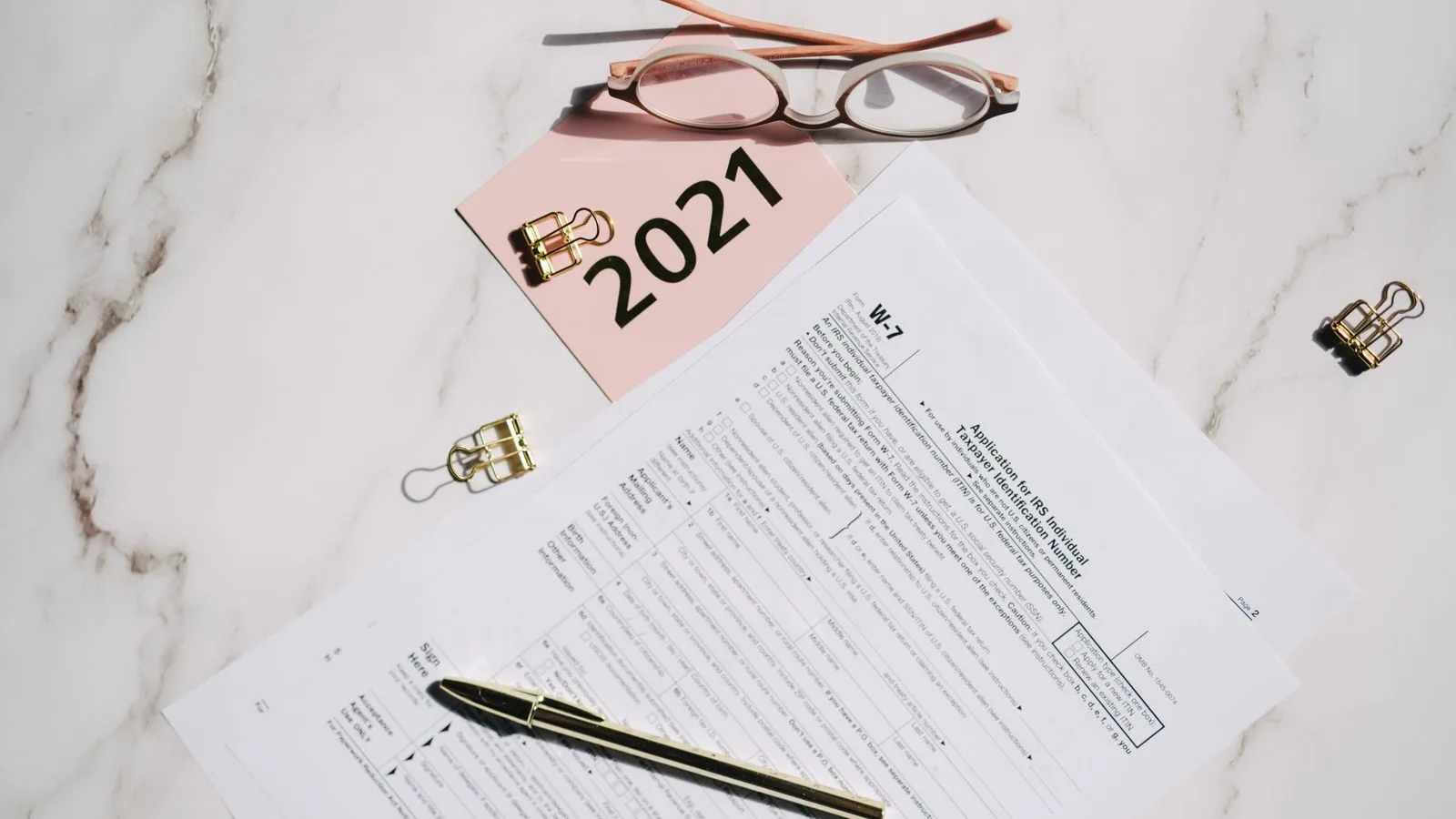 Close-up of W-7 tax forms with glasses and pen on a marble desk, ideal for finance concepts.