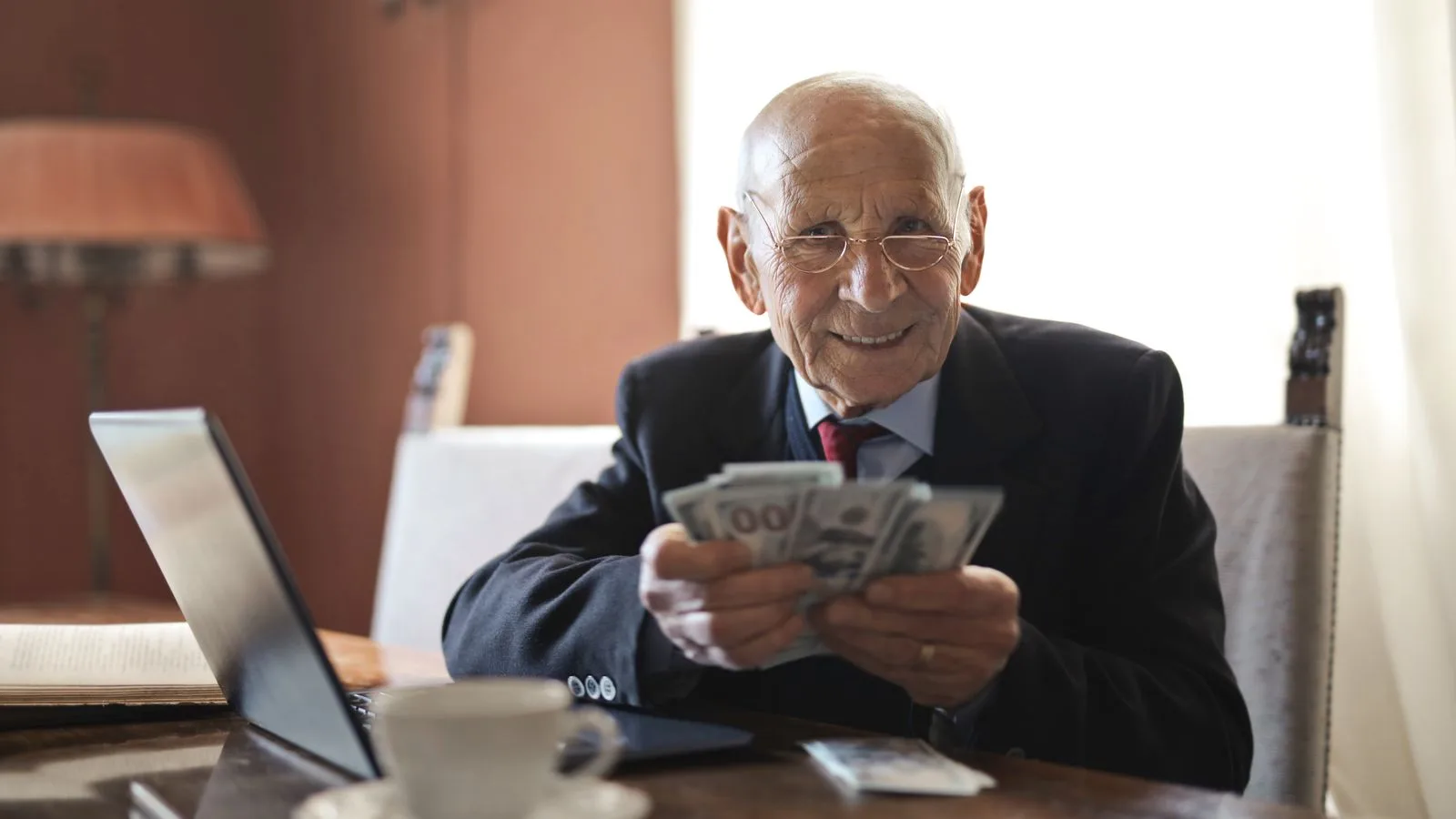 Positive senior businessman in formal suit and eyeglasses holding money in hands while sitting at table near laptop and looking at camera