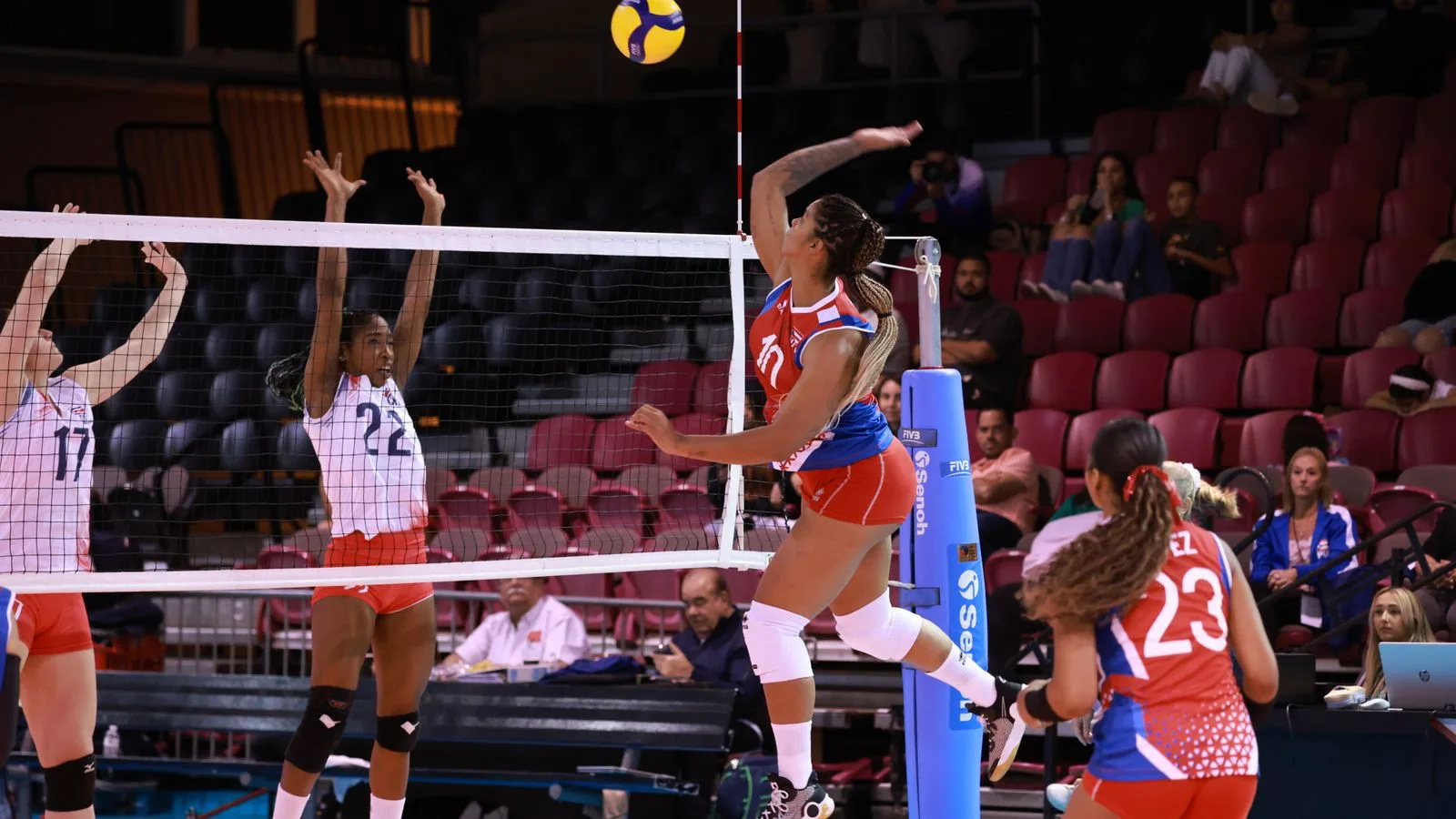 Dynamic action shot from a women's volleyball match with players in mid-air at the net.