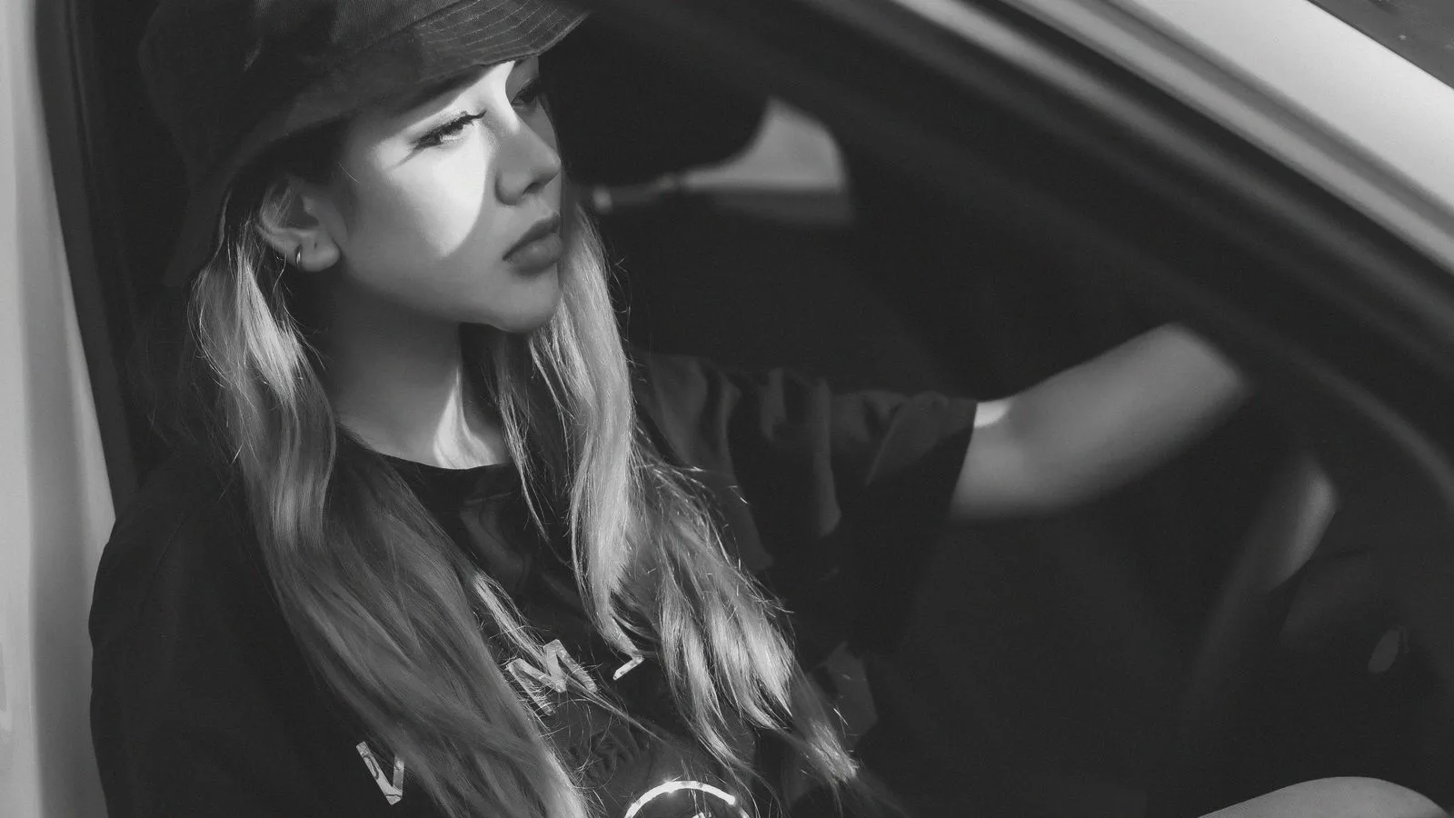 A thoughtful woman in a bucket hat sits inside a white car, captured in black and white.