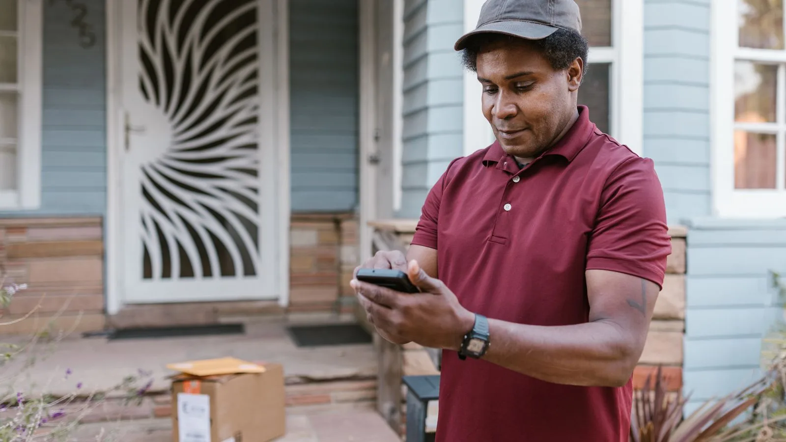 A courier in a maroon shirt checks delivery details on his phone at a residential porch with parcels.