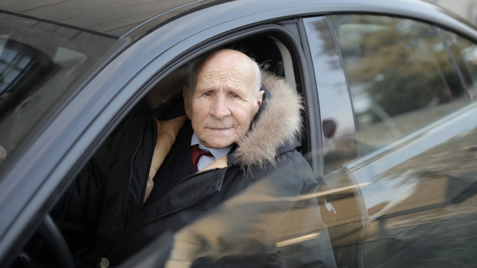 Senior man driving a car, wearing a winter coat, captured through the vehicle window.