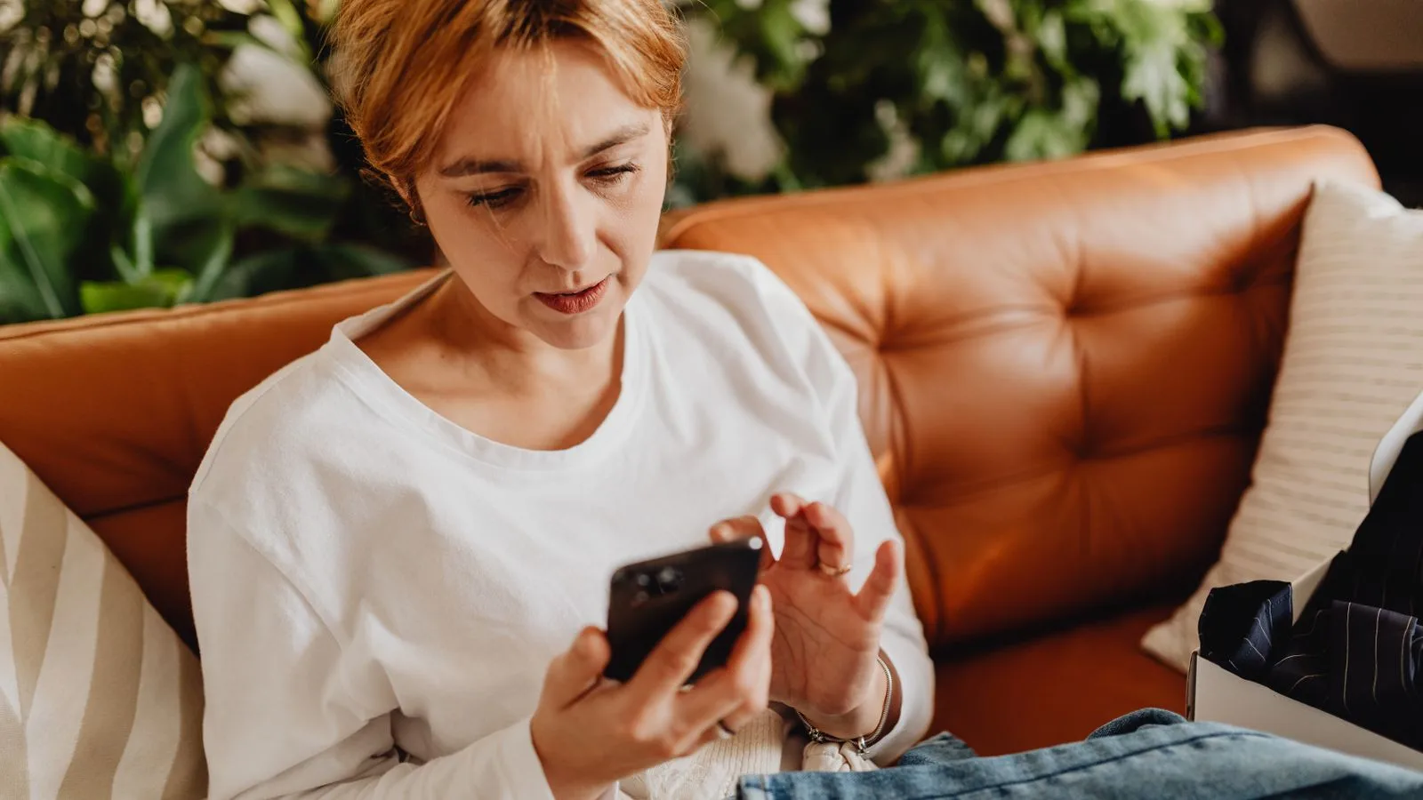 Woman in casual attire using a smartphone on a cozy sofa surrounded by plants.