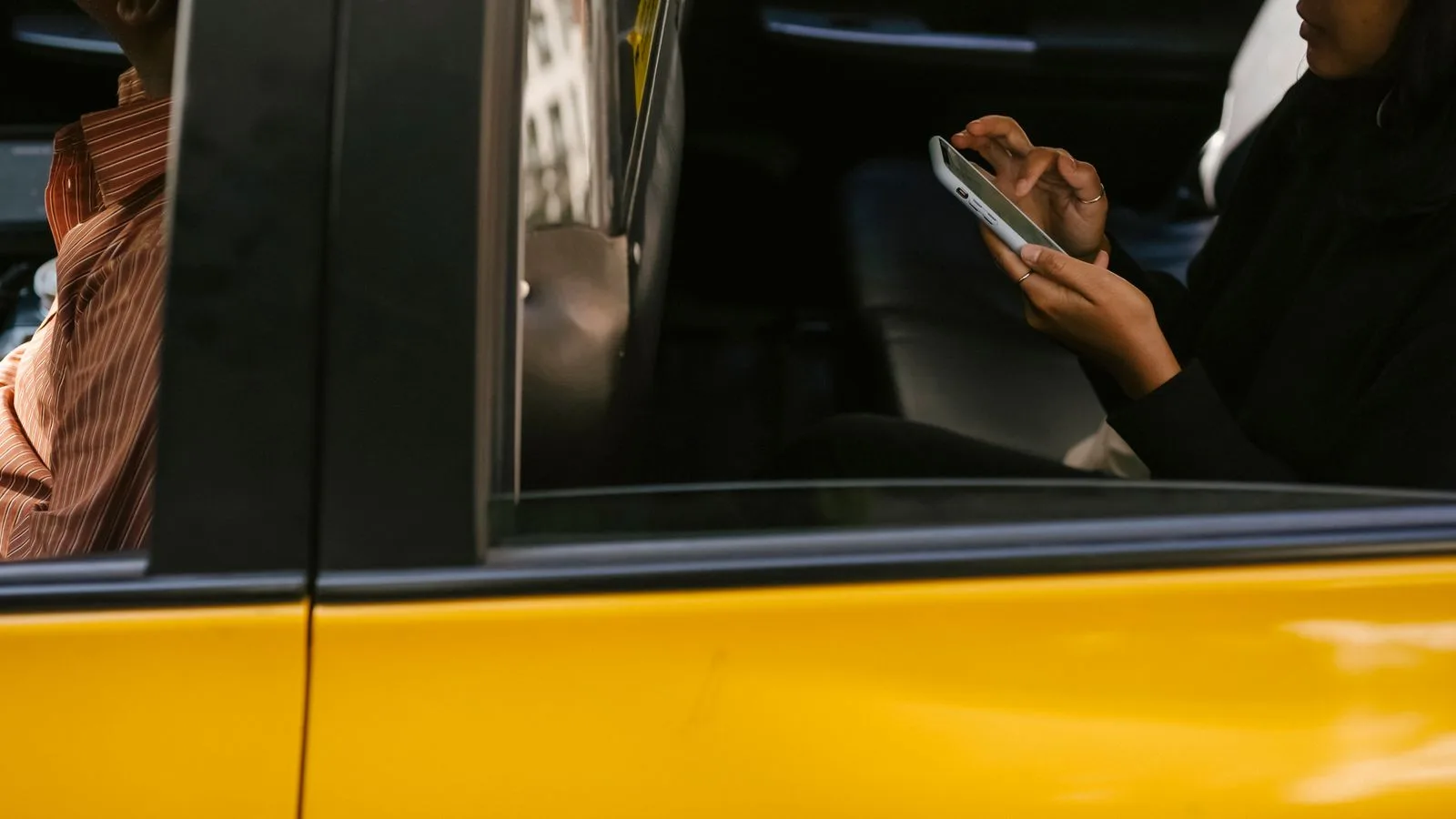 Side view of unrecognizable passenger browsing smartphone while sitting on backseat of yellow cab with anonymous driver during ride