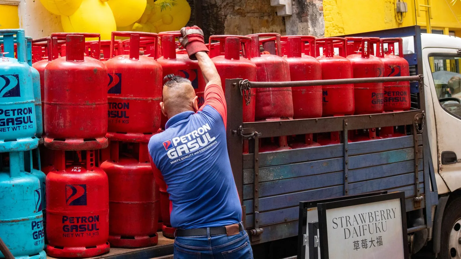 Man in uniform loading red and blue propane tanks onto a truck in urban setting.