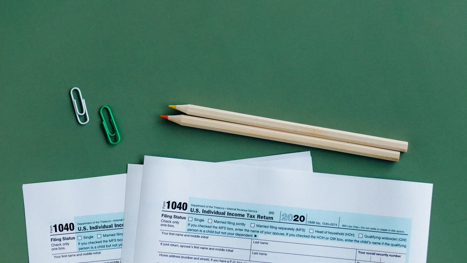 U.S. tax forms with pencils and paperclips on green surface in a flat lay setup.