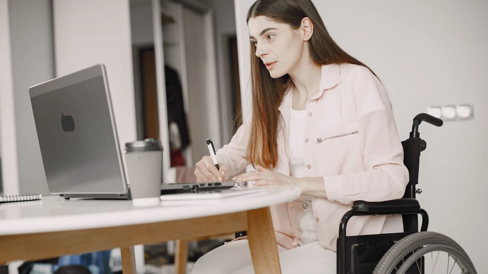 Woman in a wheelchair working remotely at home with a laptop and taking notes, showcasing remote work and accessibility.