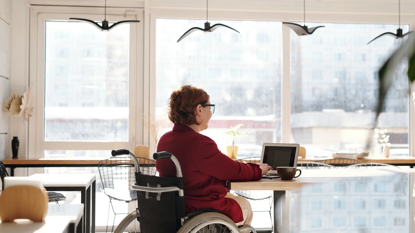 A woman in a red blazer works on a laptop in a bright café, seated in a wheelchair.