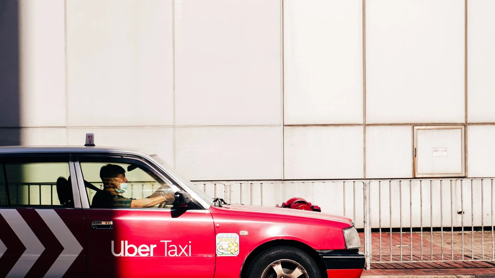 A red Uber taxi drives on a sunlit street in Hong Kong's urban landscape.