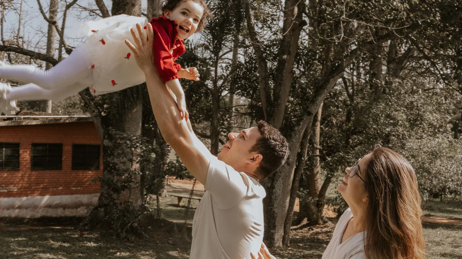 A joyful family plays in a São Paulo park, father lifting daughter high.
