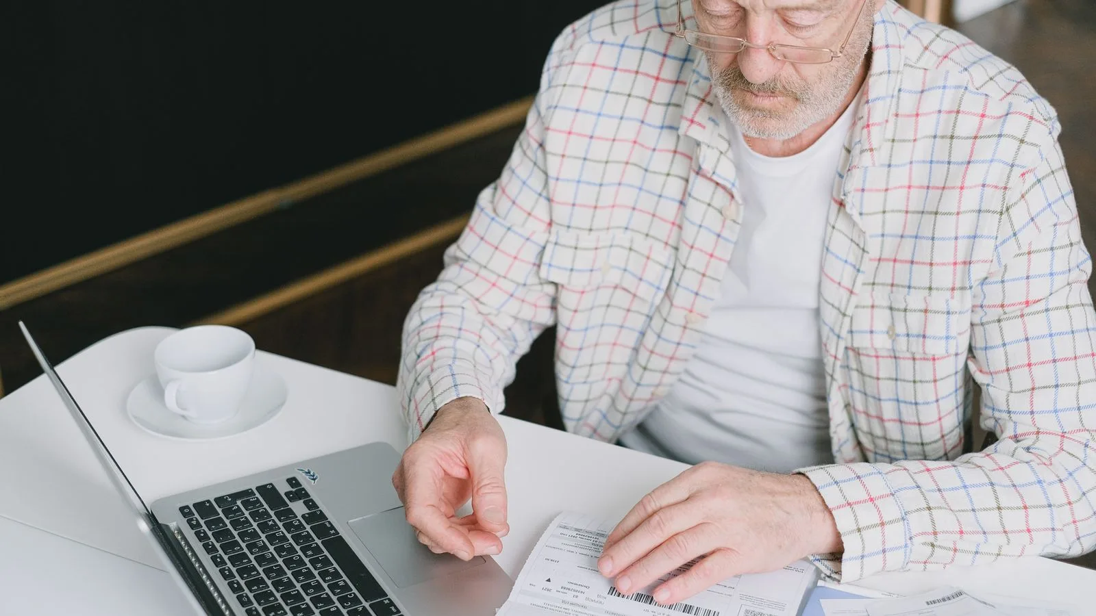 Elderly man with eyeglasses reviewing documents at a laptop. Indoor setting with natural light.