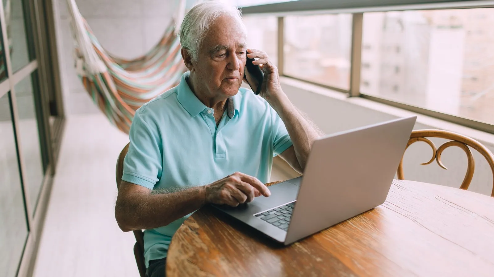 Elderly man multitasking with a phone call and laptop on a balcony.