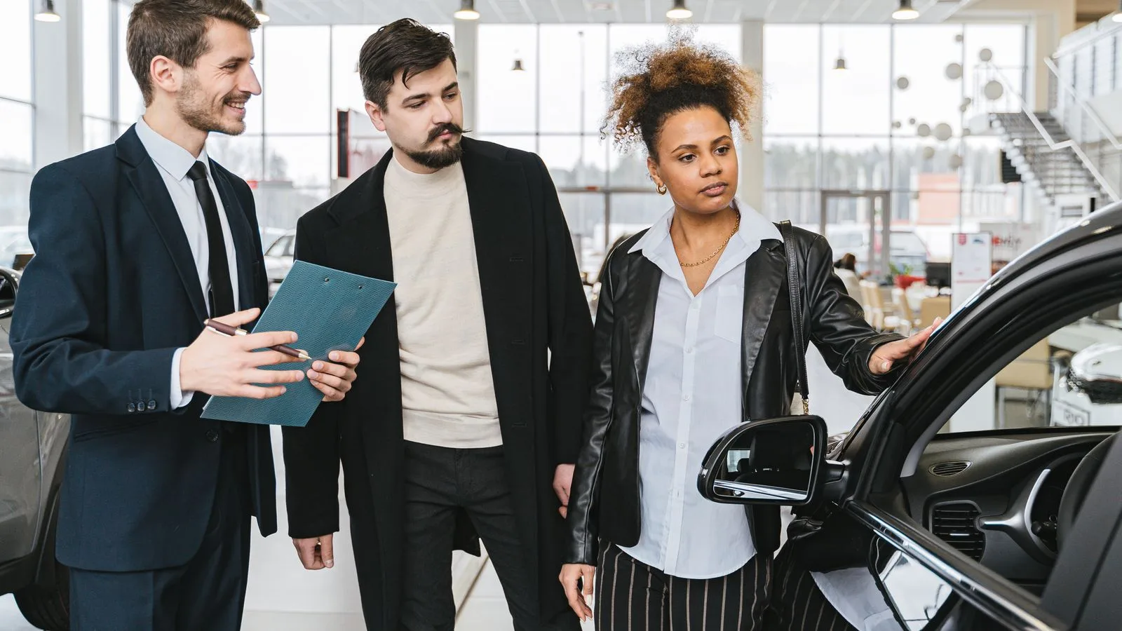 An interracial couple consults with a salesman at a car dealership, exploring vehicle options.