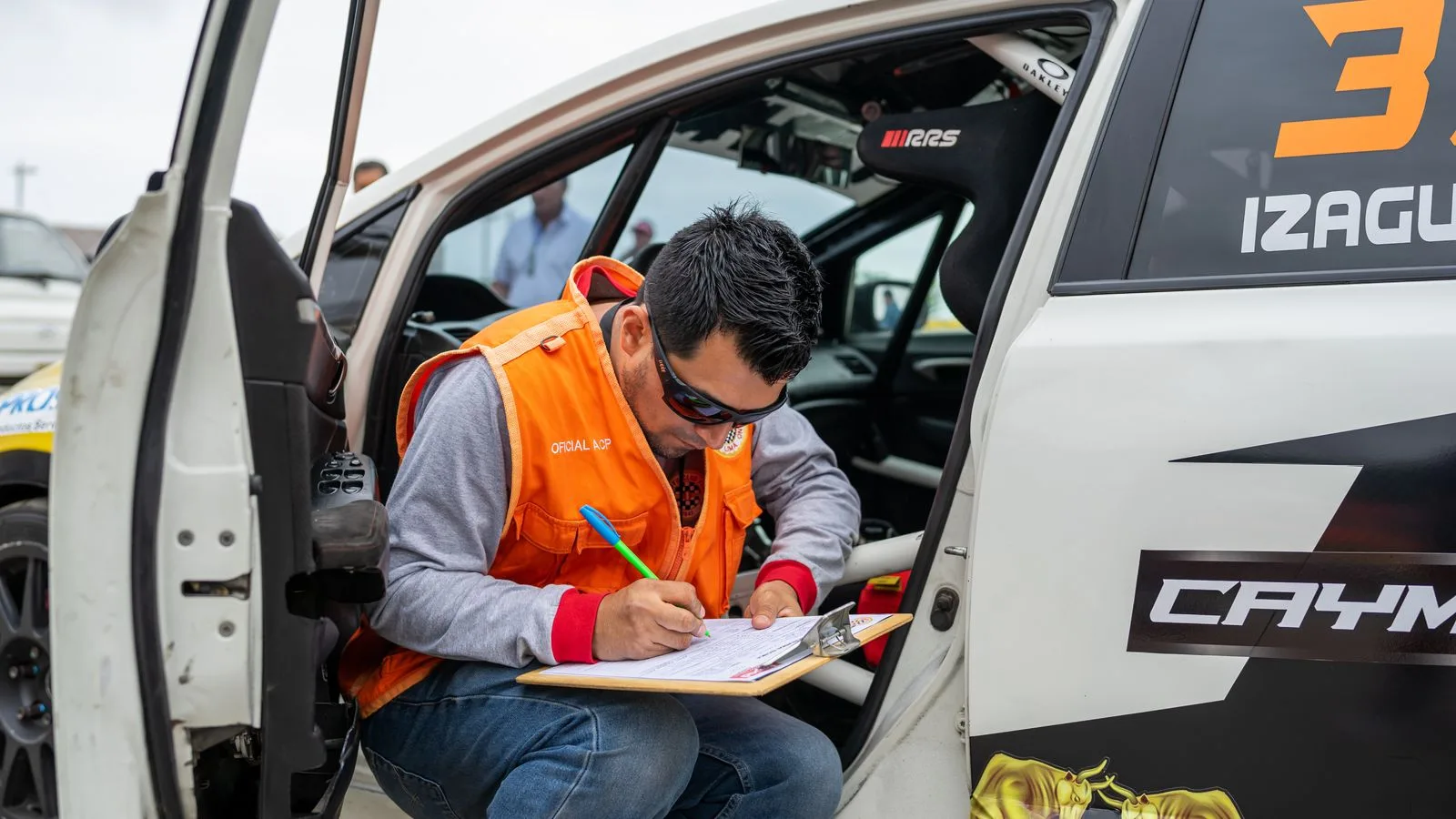 A motorsport official reviews documents while sitting by a rally car during a race event.