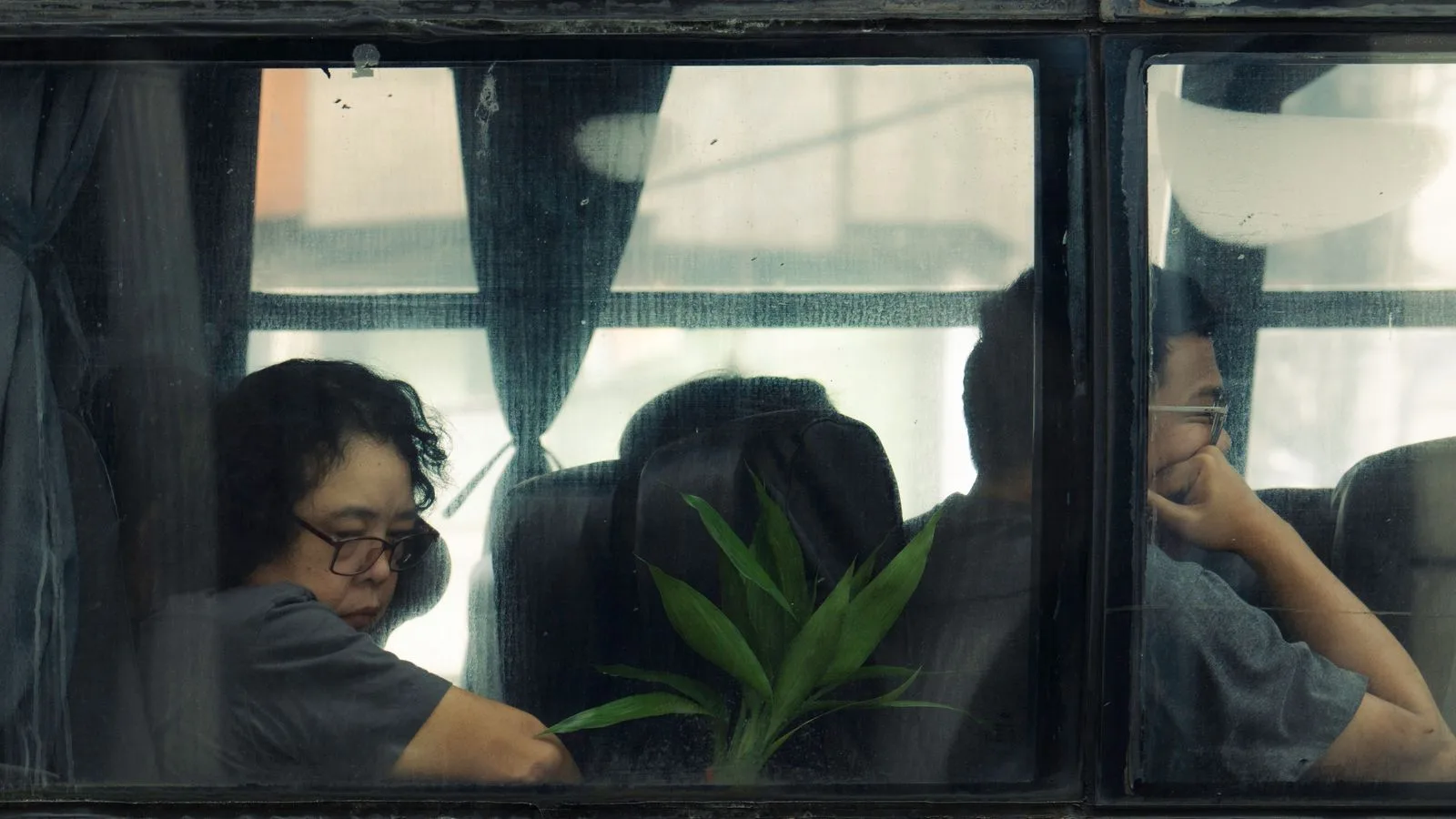 Two people sit inside a bus, viewed through a window, exuding a contemplative mood.