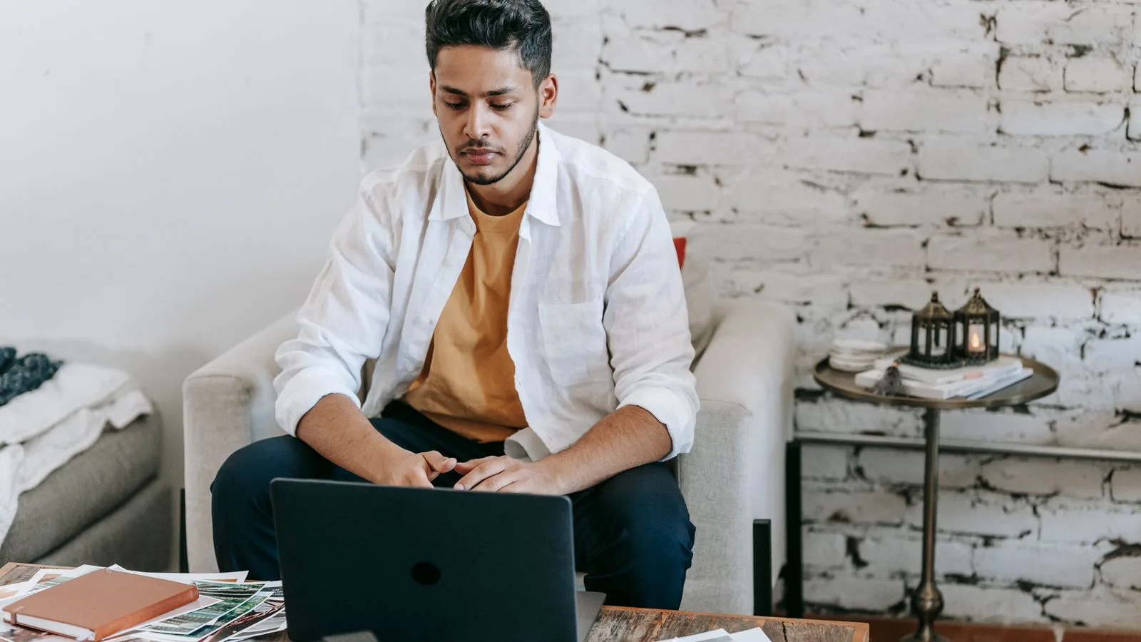 Creative young Hispanic male photographer sitting at table with photo camera and printed photos and working on laptop in modern loft style workplace at home
