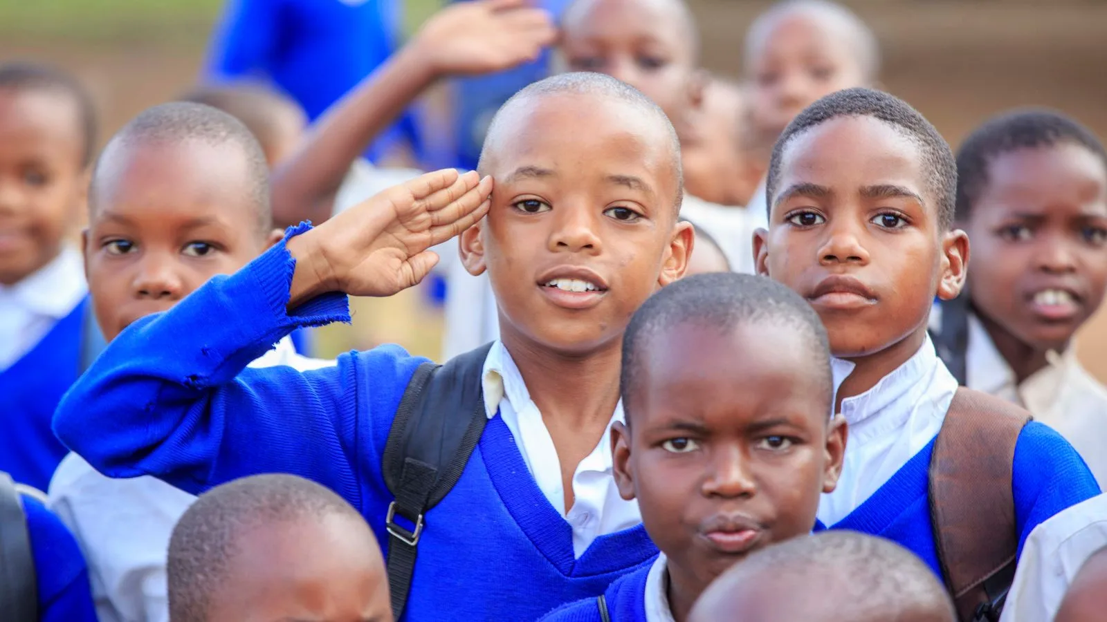 A group of school children in blue uniforms saluting outdoors on a sunny day.