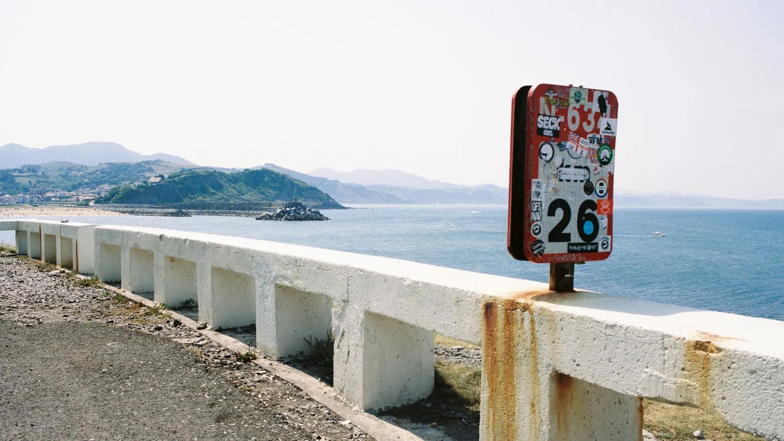 Graffiti-covered sign on a coastal road with ocean view and mountain backdrop.