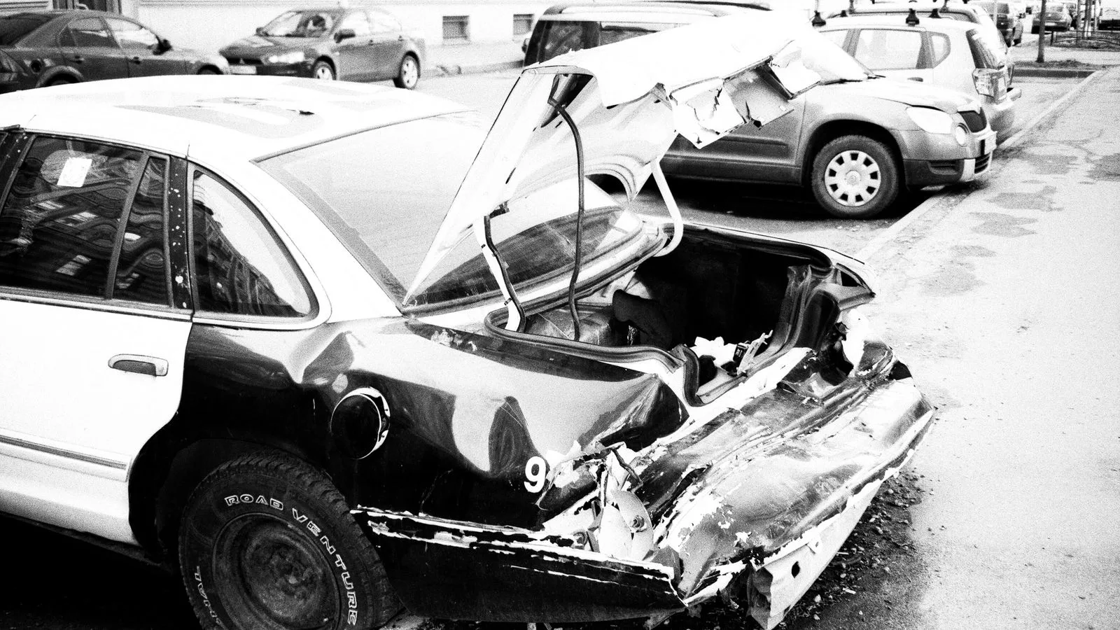 A black and white photo of a wrecked car on an urban street, highlighting vehicle damage.