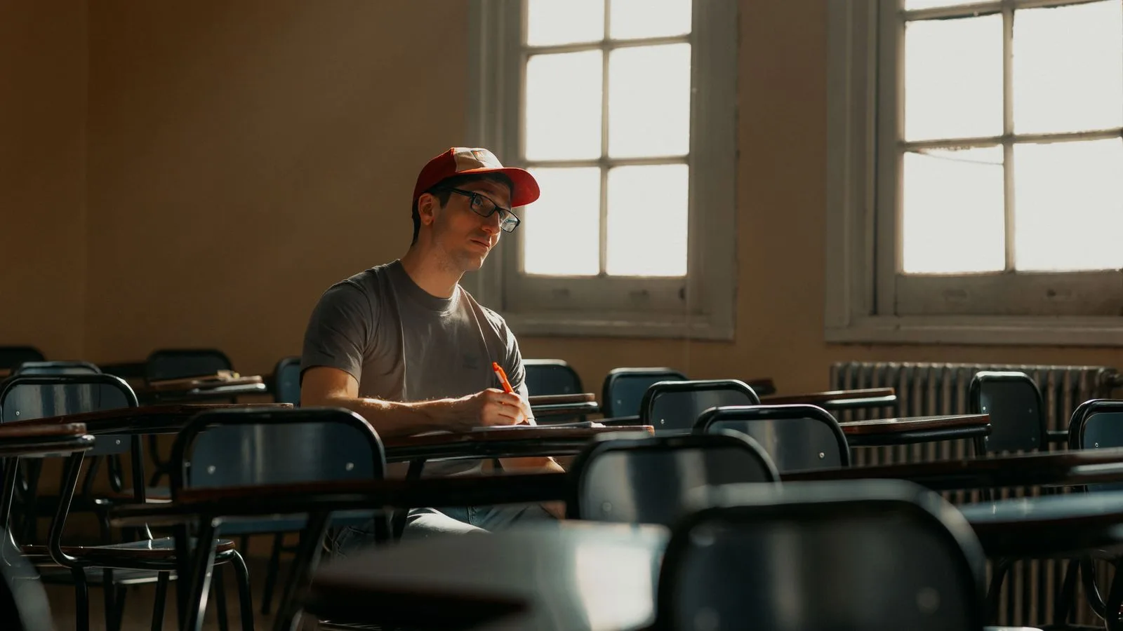 A university student in a cap studying alone in an empty classroom, lit by natural sunlight.