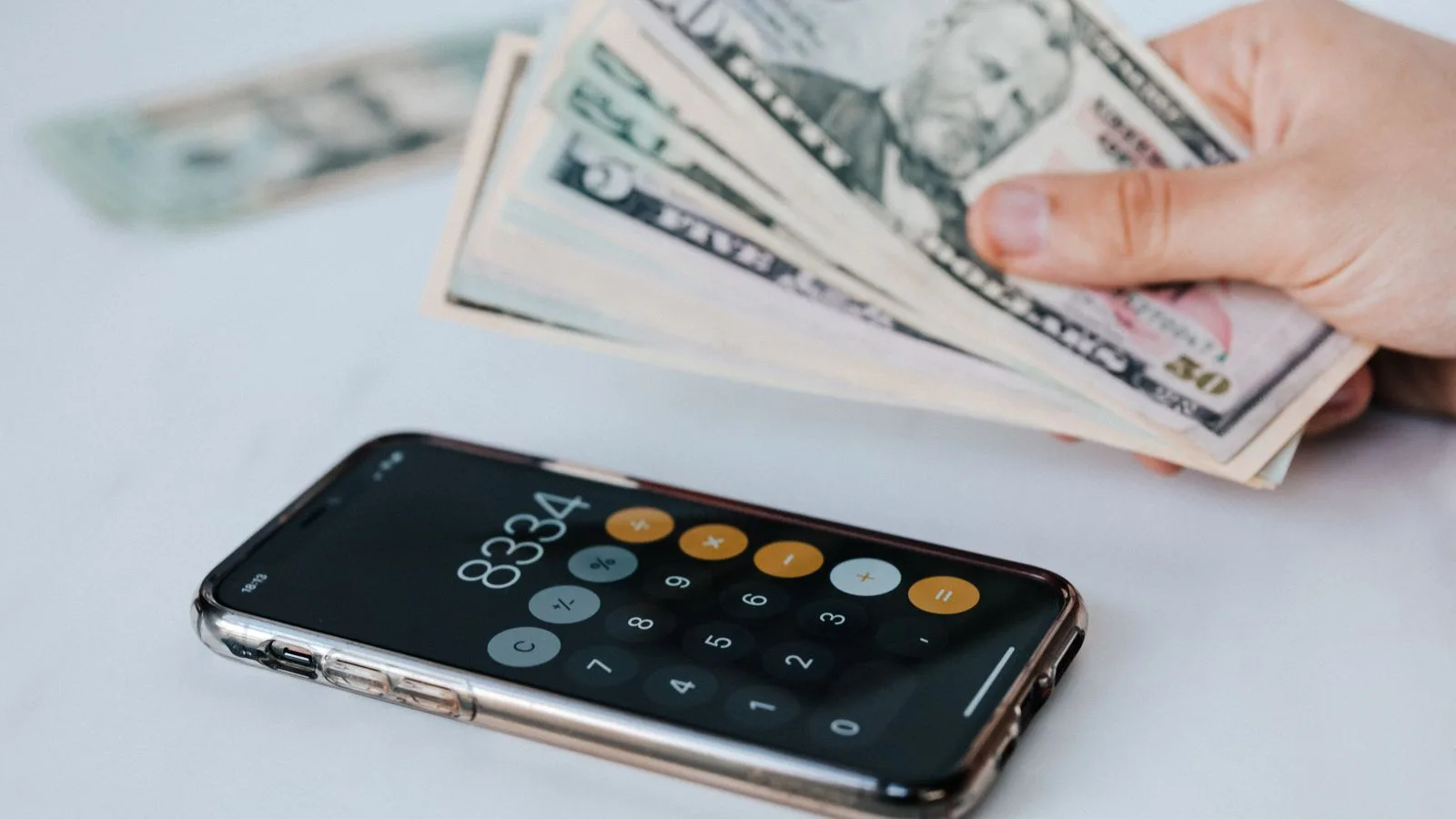Calculator application on mobile phone screen on white surface near crop anonymous person holding pile of dollars