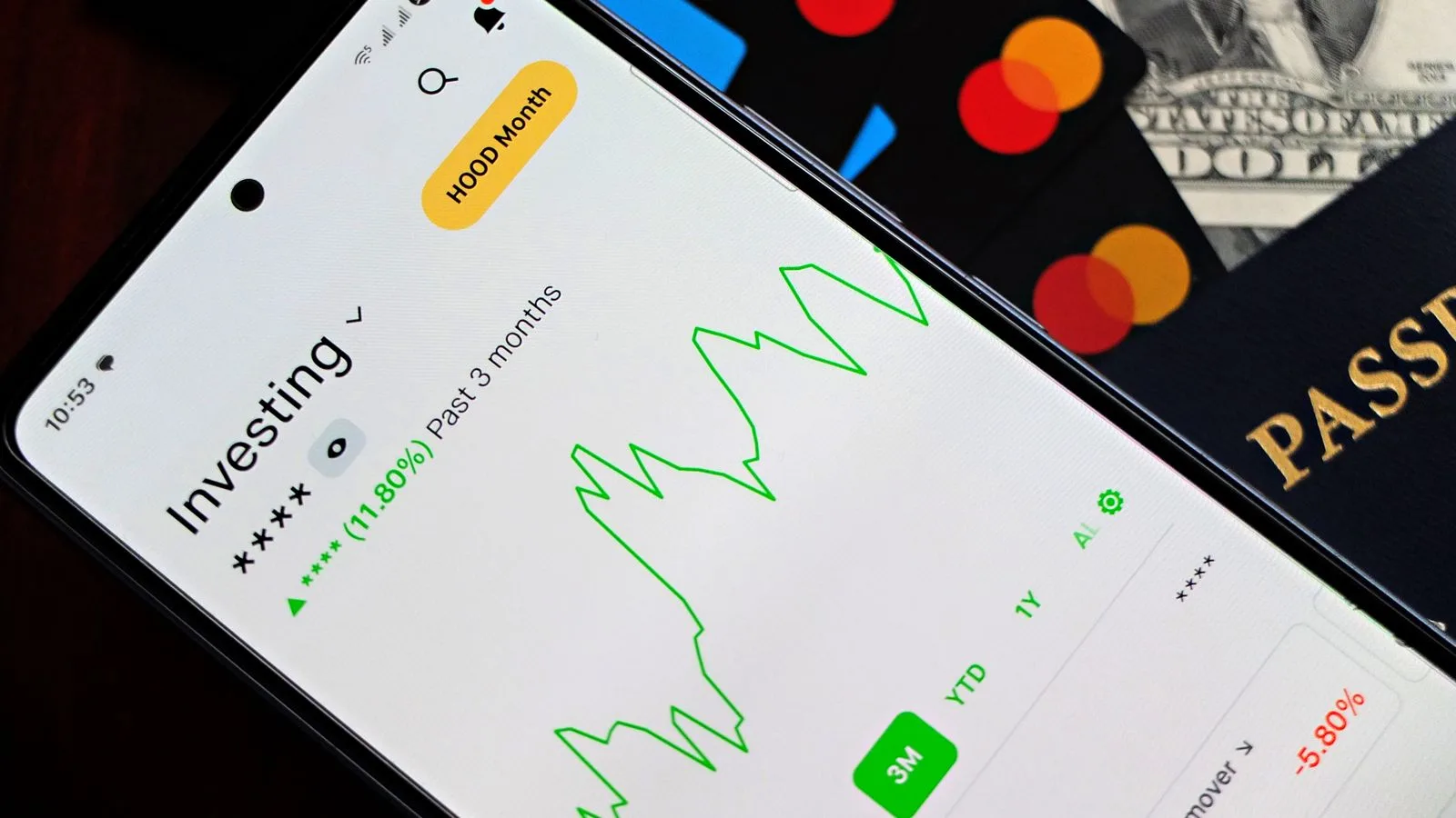 A smartphone showing an investment app with green growth indicators, surrounded by credit cards, US dollars, and a passport.