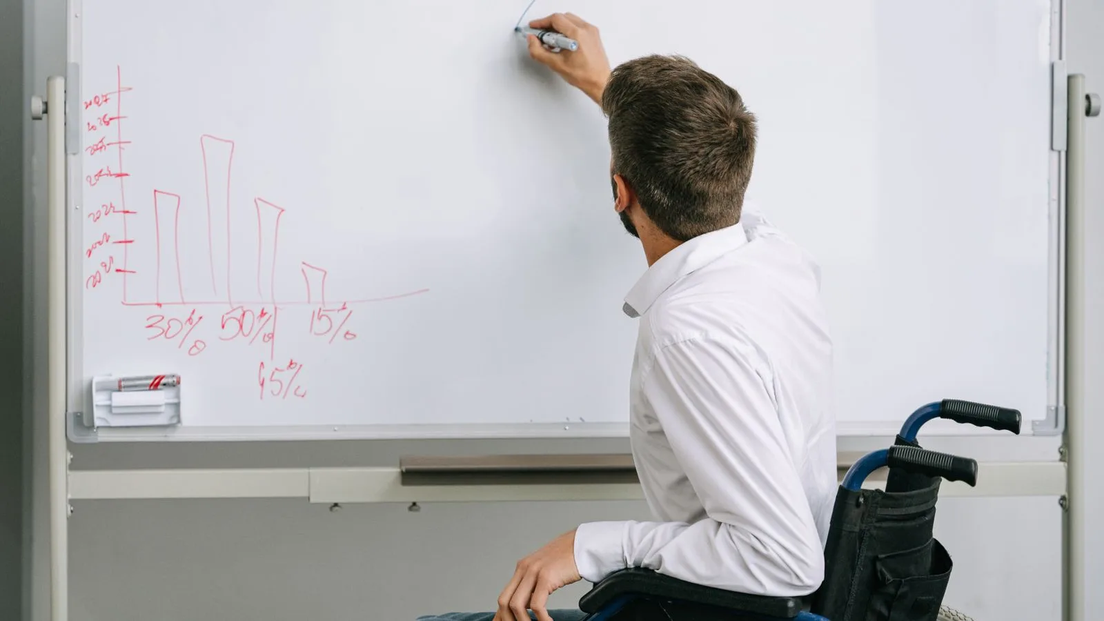 Man in a wheelchair writing on a whiteboard during an office presentation. Inclusive workplace environment.