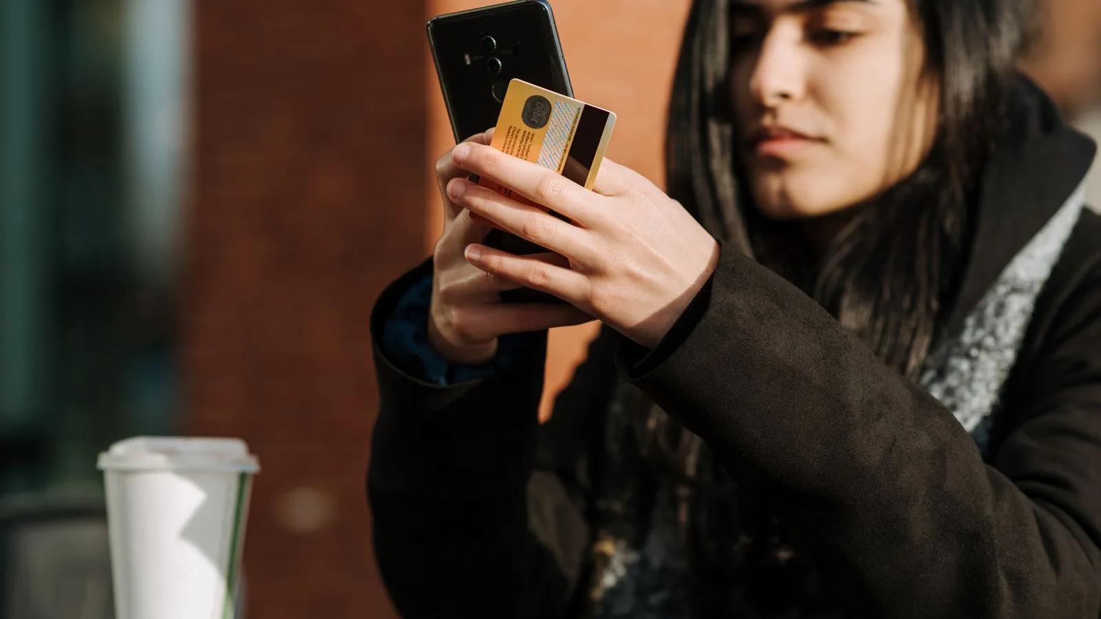 Crop young Hispanic female shopper with debit card making purchase on cellphone near takeaway beverage in cafeteria