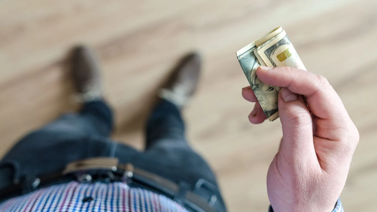 A person wearing denim and a checked shirt holds folded dollar bills in their hand indoors.