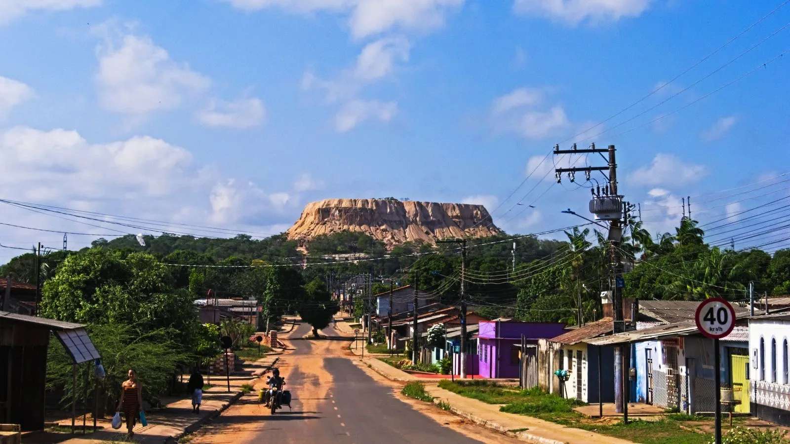 A vibrant town road with power lines leads to a majestic mountain under a blue sky.
