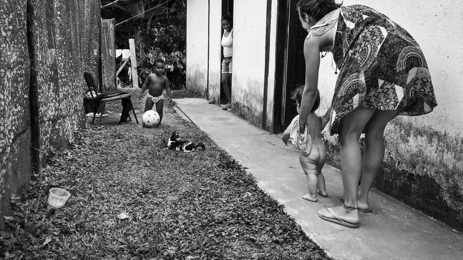 A mother helps her baby walk outside near a home while a child plays with a ball, captured in black and white.